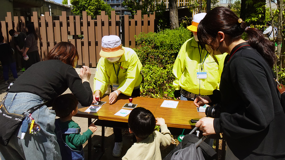 黄色いウィンドウブレイカーと、ツバが黄色くその上が白い帽子をかぶったボランティア2名が、スタンプ用紙やスタンプが置かれた机の後ろ側に立ってイベント補助をしている